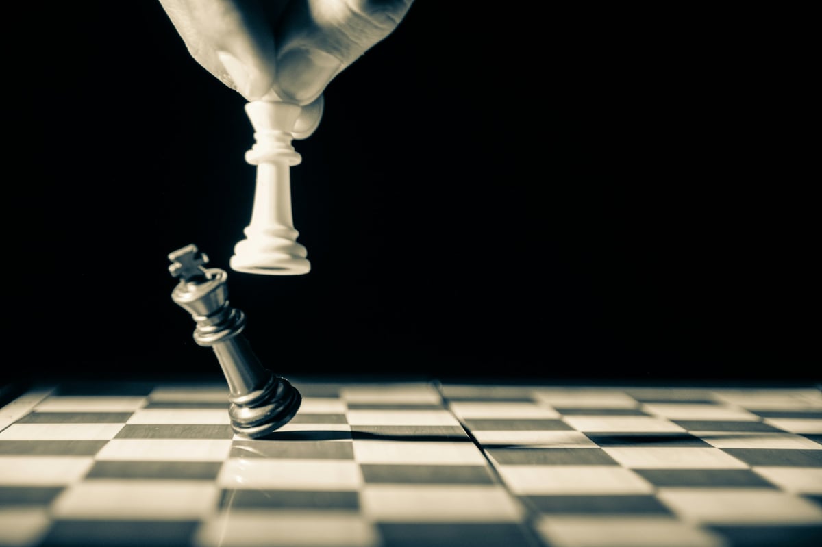 White chess piece knocking over black piece on checkered board against dark background